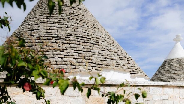 A stone building with a conical roof, a doorway with a white curtain, and potted plants. - Monte Olimpo 5 (Alberobello)