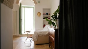 A room with a white door, a small table, a bench, a dark wooden dresser, a shelf with books and a hat, and a plant. - Monte Olimpo 5 (Alberobello)