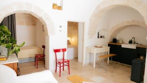 A room with a bed, a red chair, a small table, a bench, and a kitchen area with a sink and counter. - Monte Olimpo 5 (Alberobello)