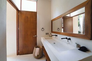 Une salle de bain avec une porte en bois, un grand miroir, un lavabo blanc et un panier en osier contenant une serviette.