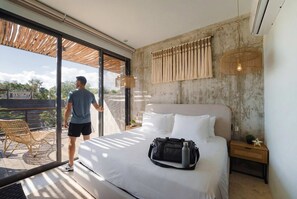A modern hotel room with a large bed, a sliding glass door to a balcony, a wicker chair, and a bedside table with a lamp. - Modern 2BR Apartment with Pool & Gym (Tulum)