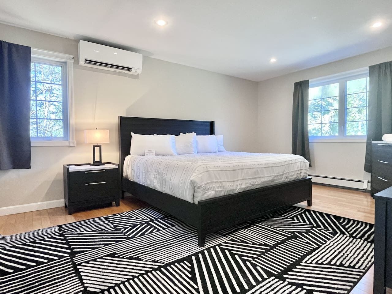 A bedroom with a bed, bedside table, lamp, air conditioning unit, and a patterned rug.