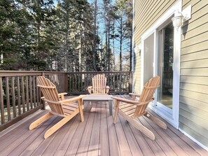 Eine Holzterrasse mit Adirondack-Stühlen und einem Tisch, umgeben von einem Geländer und einem Haus mit einem großen Fenster.