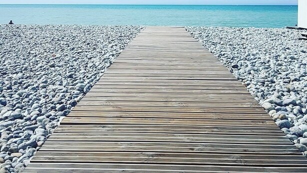 En träbrygga som leder ut till en strand med småsten och en klar himmel.