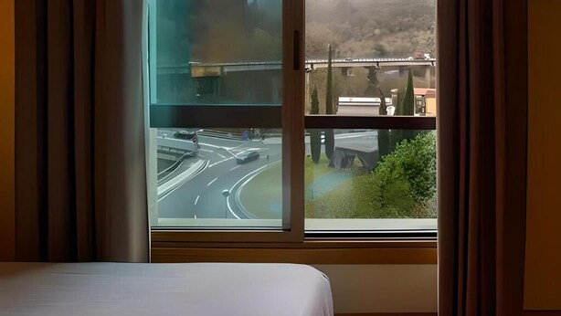 Een hotelkamer met een bed en een raam met uitzicht op een weg en groen.