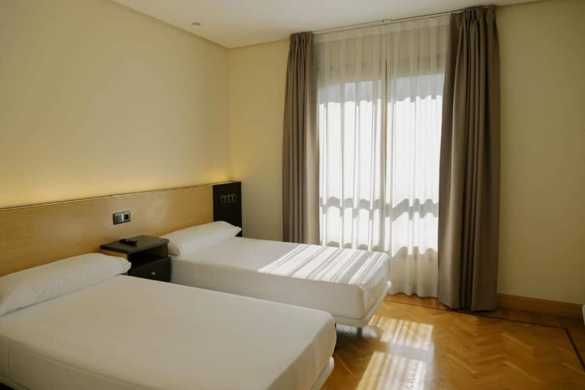 Habitación de hotel con dos camas, una mesita de noche, una ventana con cortinas y piso de madera.