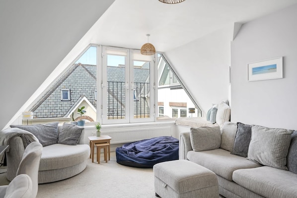 Ein modernes Schlafzimmer im Loft-Stil mit schrägverdeckter Decke, einem großen Fenster mit Blick auf benachbarte Gebäude, einer gemütlichen Sitzecke mit Sofa und Sitzsack sowie einem kleinen Tisch mit Pflanze.