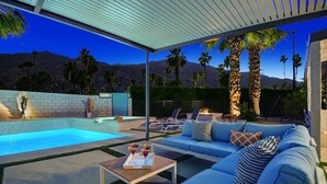 Zona de piscina con sofá, sillones, una mesa con bebidas y vistas a las montañas.