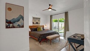 Un dormitorio con cama, escritorio, ventilador de techo, ventana con cortinas y un cuadro en la pared.