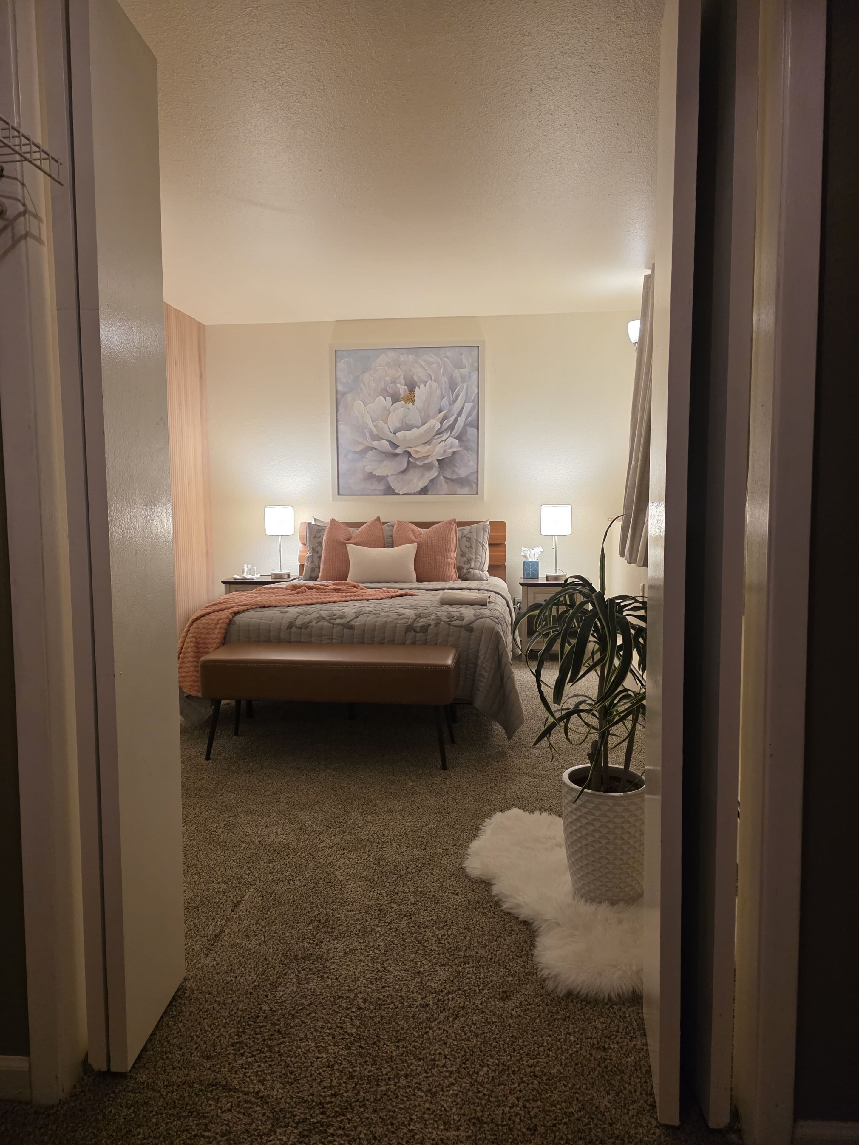 A bedroom with a bed, bedside lamps, a large floral artwork, a bench, and a potted plant.