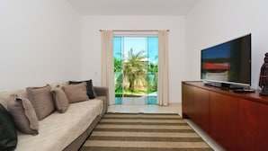 A living room with a sofa, TV, and a view through a glass door to an outdoor area. - Casa com Piscina a 250m da Praia PLH006 (Palhoça)
