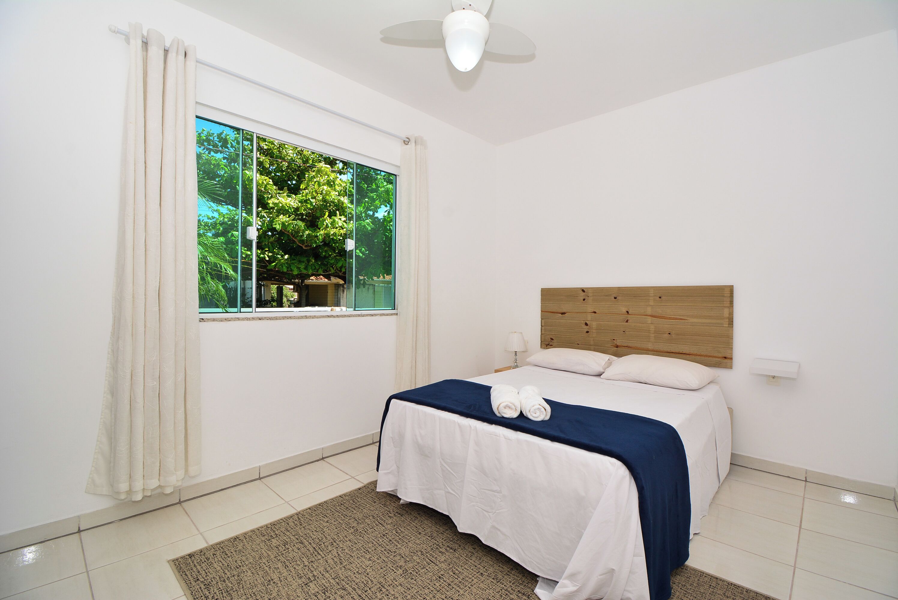 A bedroom with a bed, a window with curtains, a ceiling fan, and a view of trees outside.