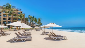 Una playa con tumbonas y sombrillas, un hotel al fondo y palmeras.