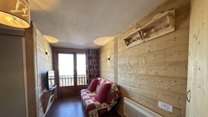 Une pièce chaleureuse avec un plancher en bois, un canapé agrémenté de coussins rouges, un téléviseur posé sur un meuble et un mur en bois orné d’une étagère décorative.