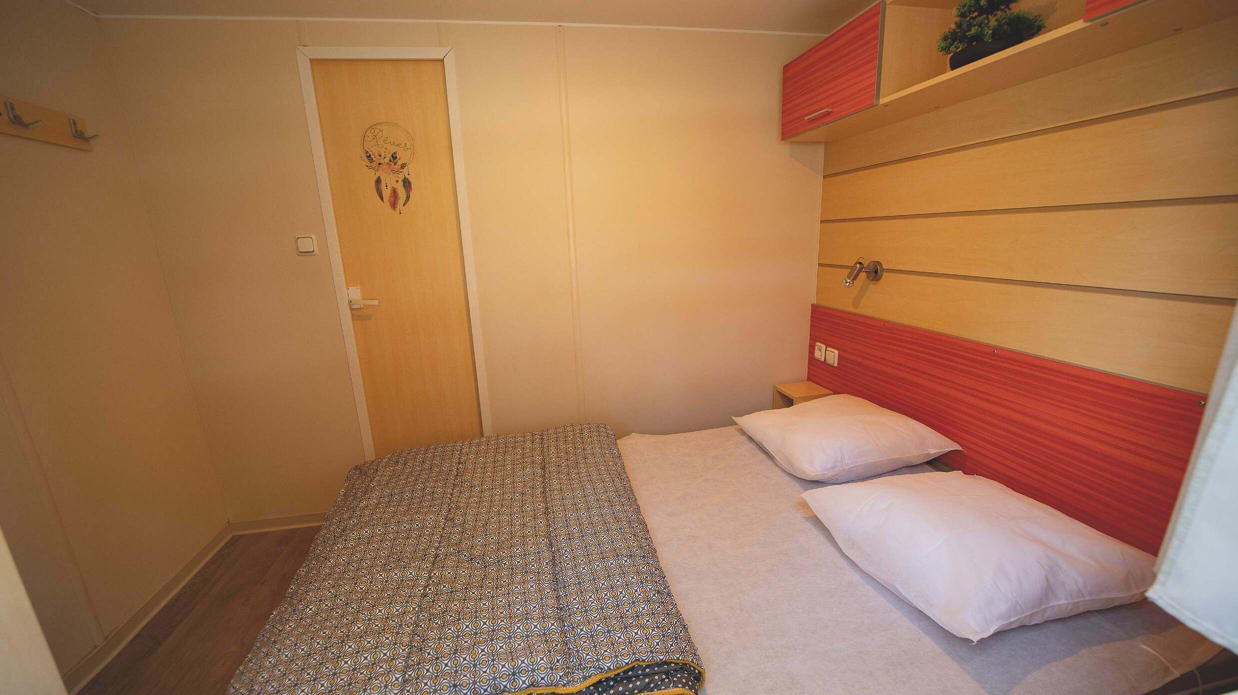 Ein kleines Zimmer mit einem Einzelbett, einer Bettdecke mit Muster, einer Tür mit Traumfänger-Dekoration und einem roten Bettkopfteil.