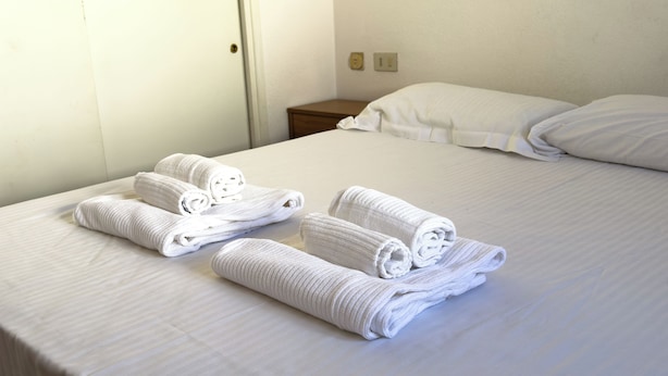 Un lit bien fait, avec des serviettes blanches pliées en rouleau et une table de chevet en bois sur laquelle se trouve une lampe.