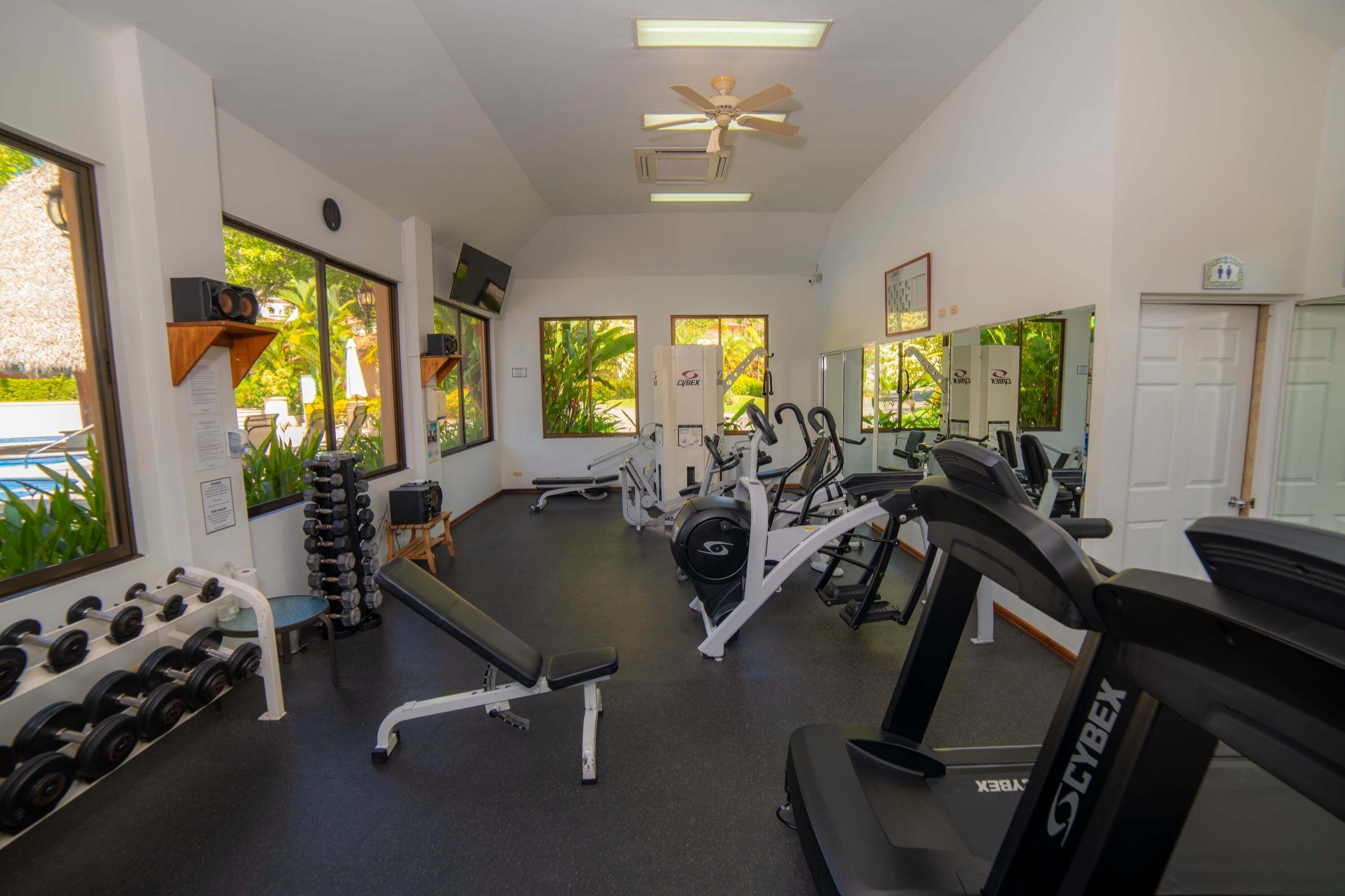 A gym with exercise equipment, a bench, dumbbells, and large windows.