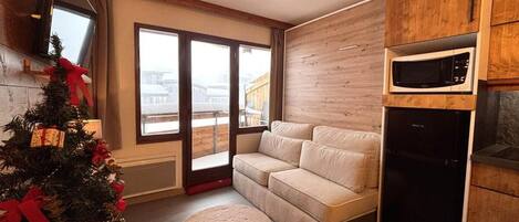 Una sala de estar acogedora con un sofá, un tapete redondo pequeño, un árbol de Navidad y una zona de cocina con microondas y refrigerador.