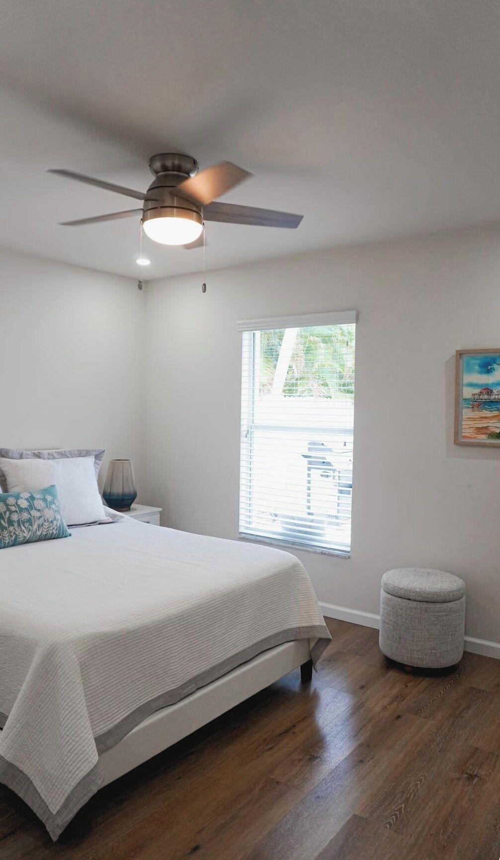 A bedroom with a bed, a ceiling fan, a window with blinds, a nightstand, and a painting on the wall.