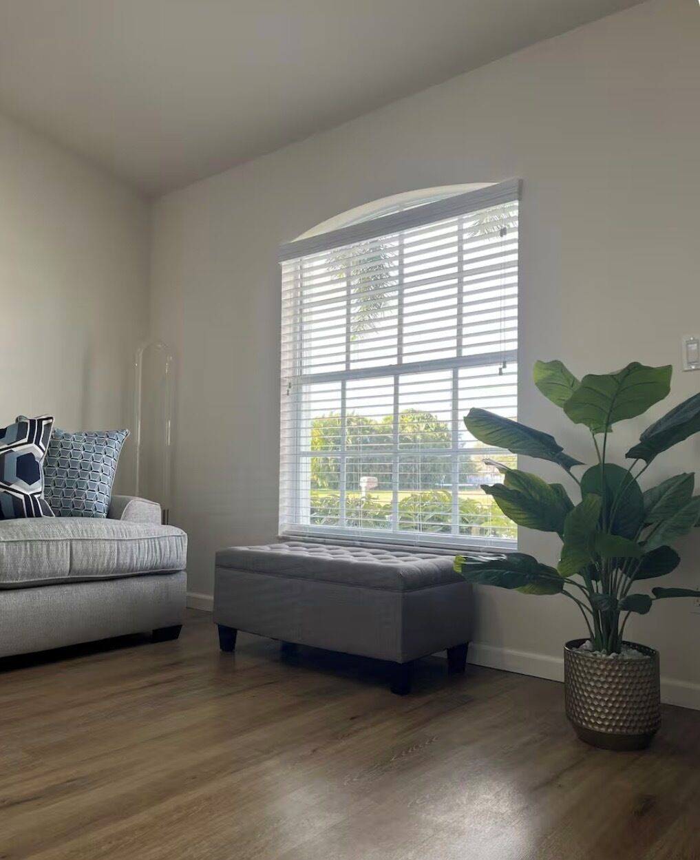 Quarto com sofá cinzento, uma janela grande com persianas, uma planta em vaso e piso de madeira.