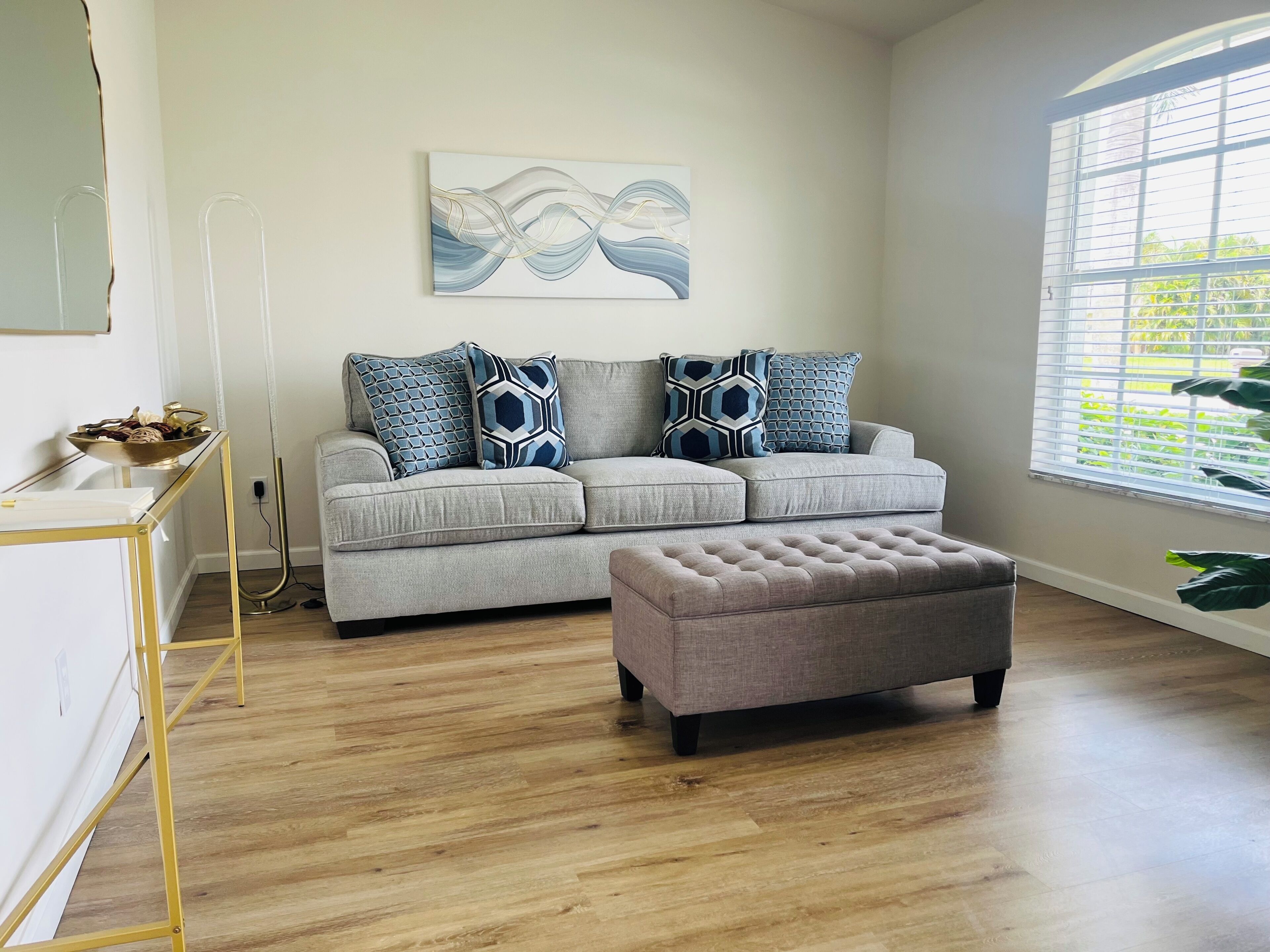 A living room with a grey sofa, matching ottoman, and a painting on the wall.