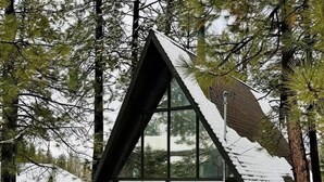 Eine dreieckige Holzhütte mit großem Fenster, einer Holzterrasse und einem mit Schnee bedeckten Dach.