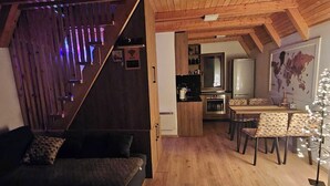 A cozy living area with a wooden staircase, a sofa, a dining table with chairs, and a kitchen in the background.