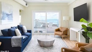 A living room with a sofa, armchair, coffee table, and a TV on the wall. -  Atlantic's Edge @ Carolina Beach—Panoramic views of the ocean + pool /hot tub (Carolina Beach)