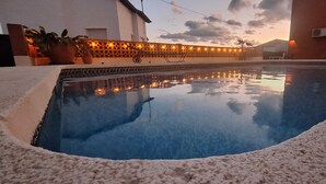 Ein Swimmingpool mit einer gefliesten Kante, umgeben von einer weißen, strukturierten Fläche und einem dekorativen Zaun mit Beleuchtung im Hintergrund.
