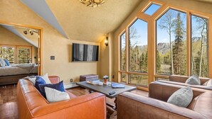 Ein geräumiges Wohnzimmer mit Ledersofas, einem Couchtisch und einem großen Fenster mit Blick auf Bäume und Berge.