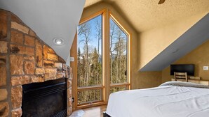 Ein Schlafzimmer mit Kamin aus Stein, großen Fenstern mit Blick auf einen Wald und einem Bett.