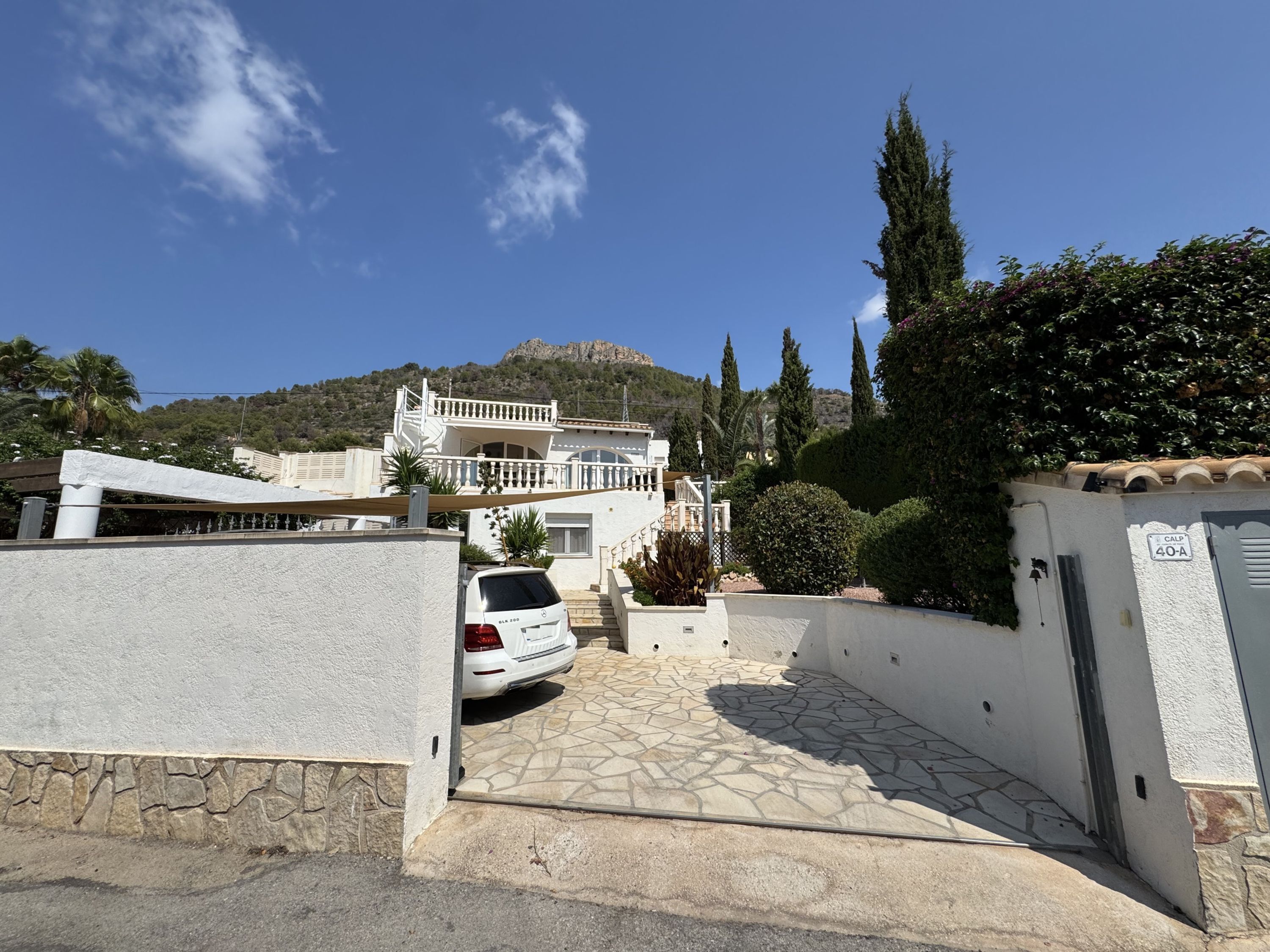 A residential building with a stone-paved driveway, a white car parked in front, and a mountain in the background.