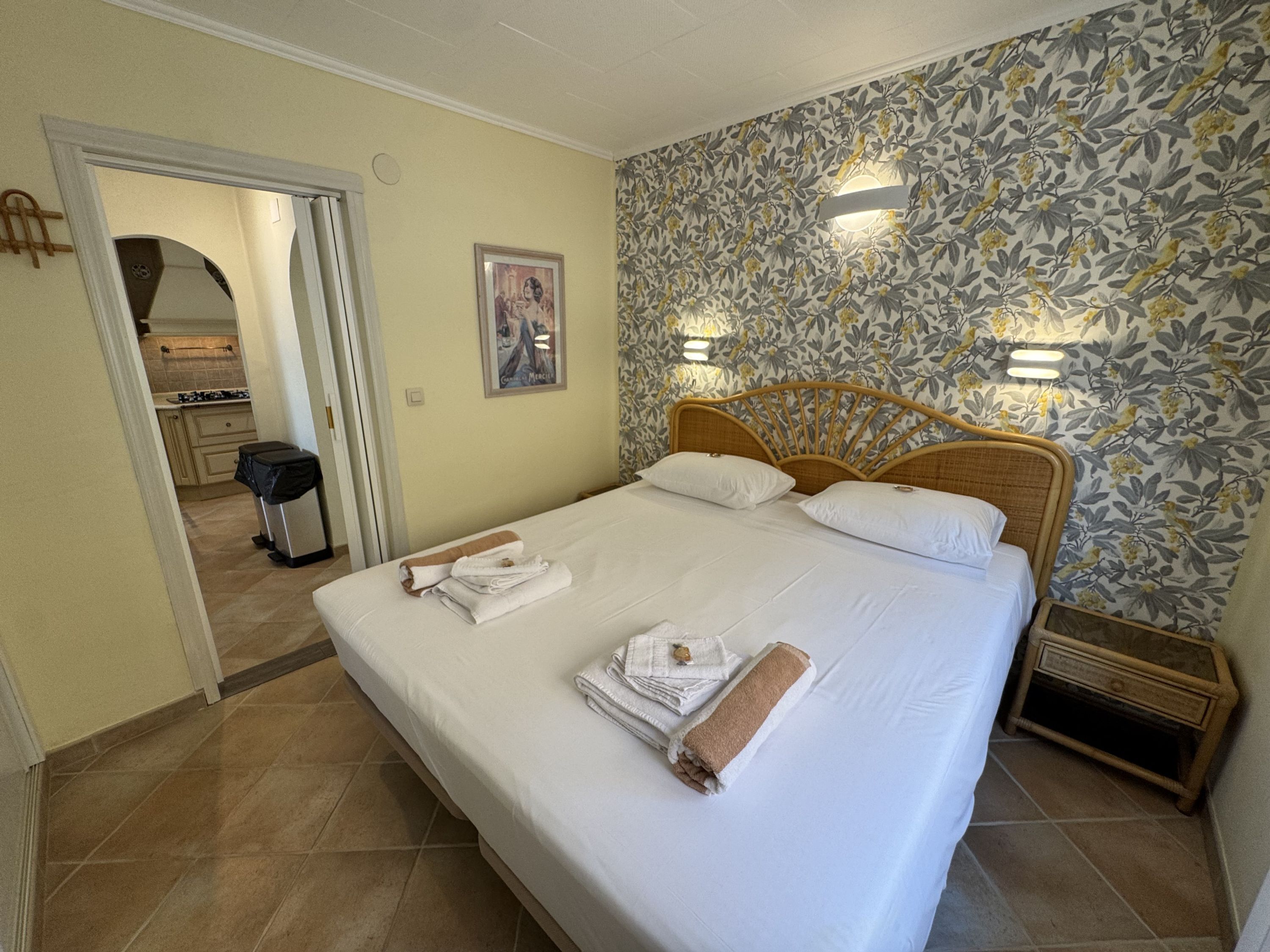 A hotel room with a bed, towels, a wooden headboard, a floral wallpaper, and a small bedside table.