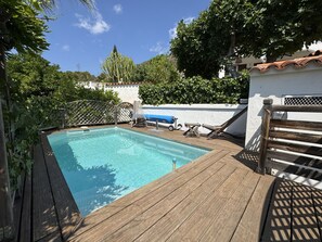 Ett trädäck med en swimmingpool, omgivet av grönska och en vit vägg.