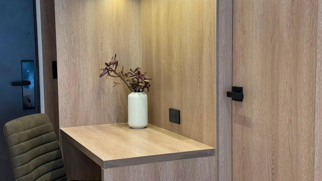 Un bureau en bois sur lequel se trouve un vase blanc contenant des fleurs séchées, une prise électrique noire et une chaise verte.