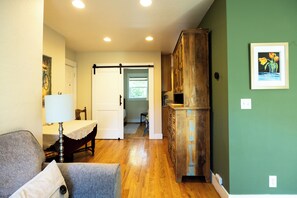 A room with a wooden cabinet, a table with a lamp, a chair, and a door leading to another room. - The Hideaway Cottage at Second Street Cottages (Ashland)