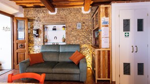 Ein gemütliches Zimmer mit Steinwand, Holzbalkendecke, einer Couch, einem Sessel, einer Kommode mit Unterlagen und einer Tür mit Erste-Hilfe-Ausrüstung.