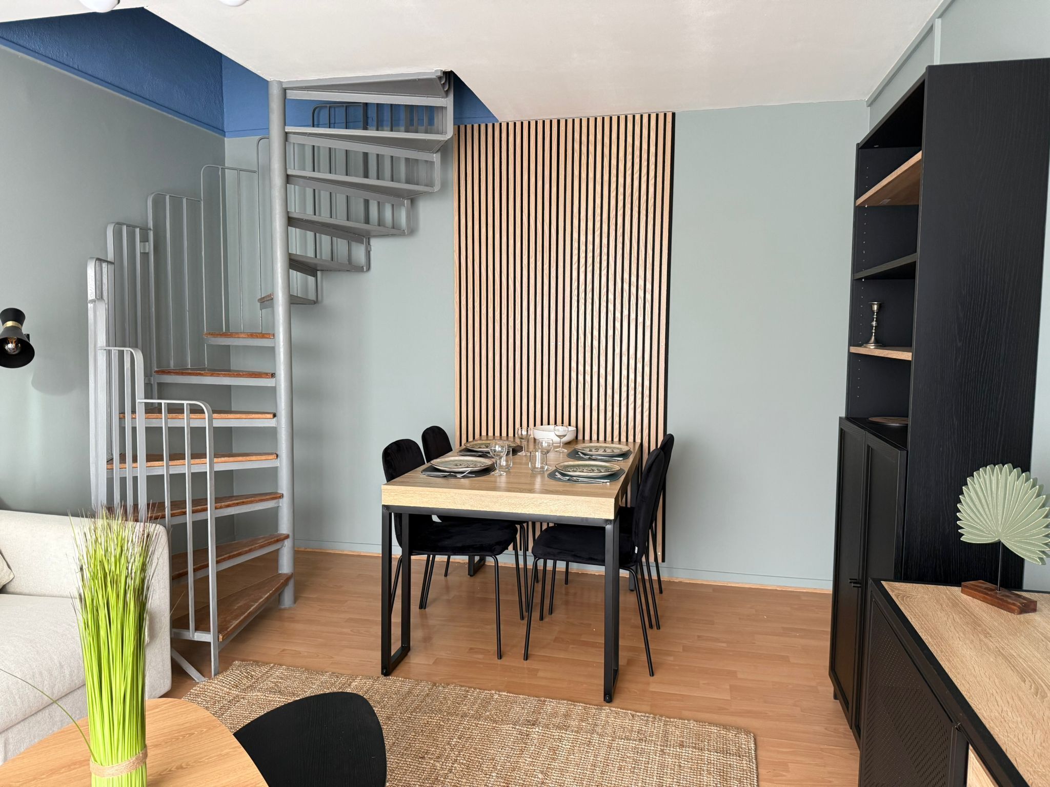 Un espace repas avec une table en bois, des chaises noires et un mur composé de lattes de bois disposées verticalement.
