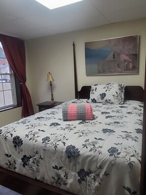 A bedroom with a bed, a nightstand, a lamp, a window with curtains, and a painting on the wall.