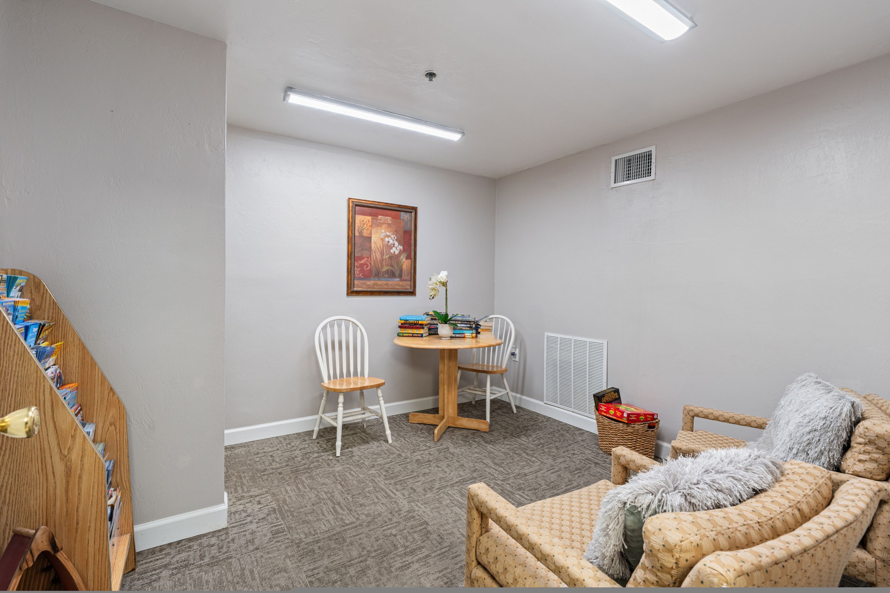 A room with a wooden desk, two white chairs, a round table with books and a vase, a wicker chair, and a grey sofa.