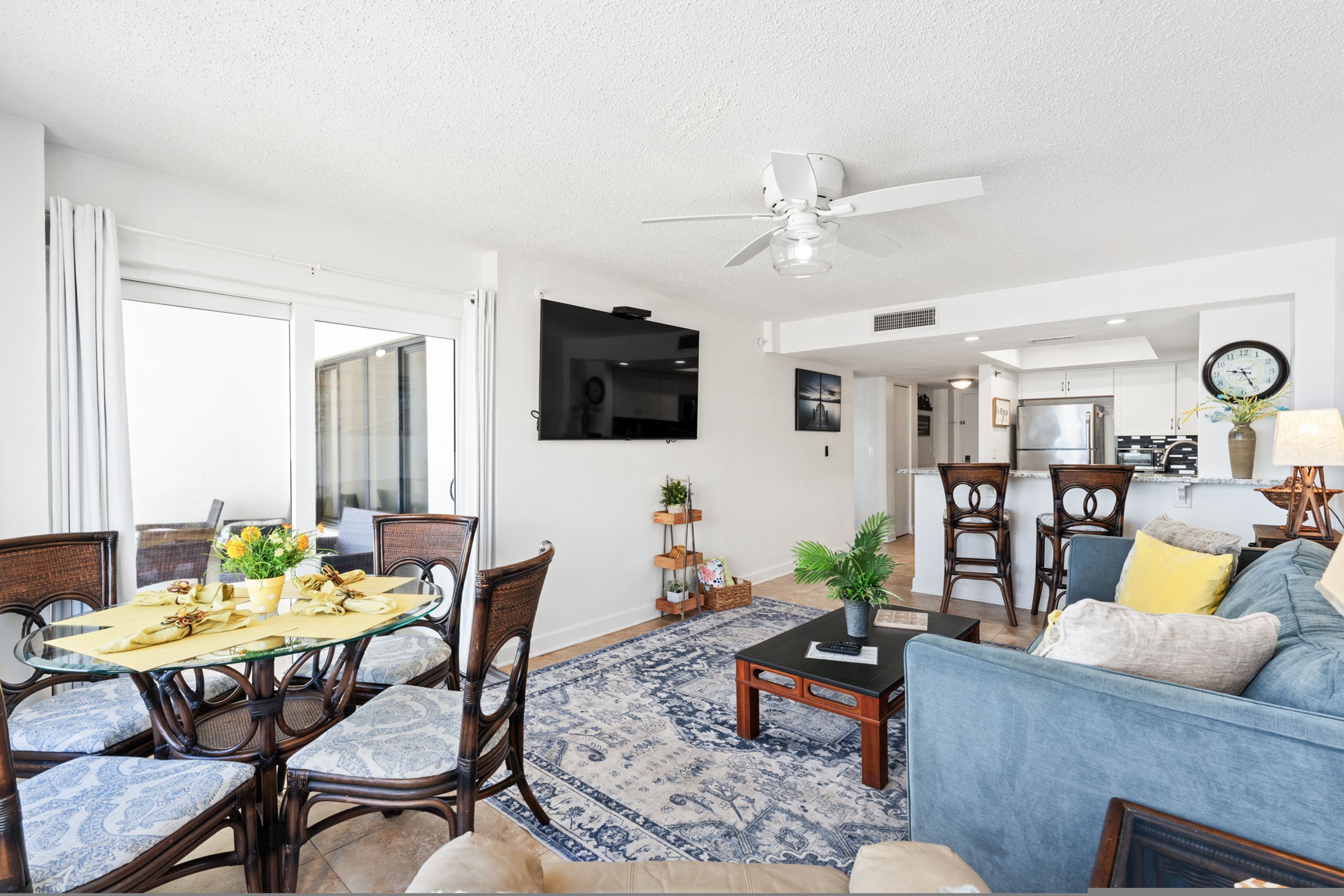 A living room with a glass dining table, blue patterned chairs, a blue sofa, a coffee table, and a ceiling fan.
