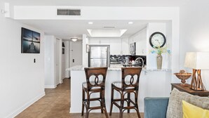 Una cocina moderna con armarios blancos, un refrigerador de acero inoxidable y un horno empotrado. Hay dos sillas de mimbre en una pequeña isla con encimera de mármol.