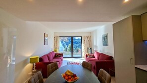 Ein Wohnzimmer mit Esstisch, zwei roten Sofas, einem Fernseher und einem großen Fenster mit Blick auf Bäume.