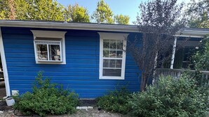 Une maison bleue avec deux fenêtres, un petit jardin avec de la pelouse et des arbustes, et un robinet extérieur bien visible.