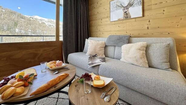 Een gezellige kamer met een grijze bank, een houten tafel met eten, en een raam met uitzicht op de bergen.