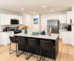 A modern kitchen with white cabinetry, a central island with bar stools, stainless steel appliances, and a decorative vase with flowers. - Near Navarre Beach- Sauna, Hot Tub, Arcade Game Room, Sunsets (Navarre)