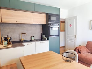 Eine kleine Küche mit Mikrowelle, Backofen und Spüle. Ein Holztisch und ein Stuhl mit Drahtkorb sind vorhanden. Eine Tür mit einer Nummer ist sichtbar.