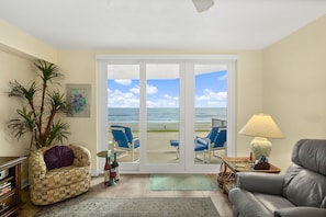A living room with a view of the beach, a sofa, a chair, a lamp, and a bookshelf. - Oceanfront 2BR Condo Ponce Inlet Sleeps 6 (Ponce Inlet)