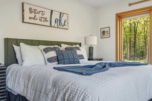 A neatly made bed with a white and blue patterned bedspread, a decorative pillow, and a blue knitted throw. A framed sign with the text 'Better to wake at the lake' is on the wall.