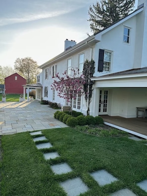 Une maison blanche avec un chemin de pierre menant à l’entrée, entourée de verdure et d’un petit arbre aux fleurs roses.